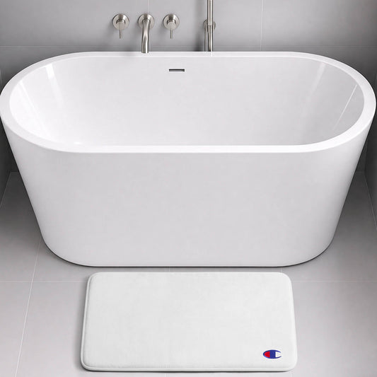 A modern white freestanding bathtub with a wall-mounted faucet sits on gray tile. In front of the tub is the Champion Logo Patch Memory Foam Bath Mat—a white, rectangular bath mat crafted from memory foam for extra comfort and designed to be skid-resistant. A small blue and red Champion logo patch adds a subtle yet stylish touch.