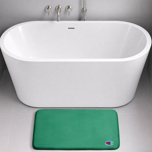 A modern white bathtub with wall-mounted fixtures sits on a gray tile floor. In front of the tub is a green Logo Patch Memory Foam Bath Mat from Champion, featuring a small, colorful C logo in the bottom right corner.