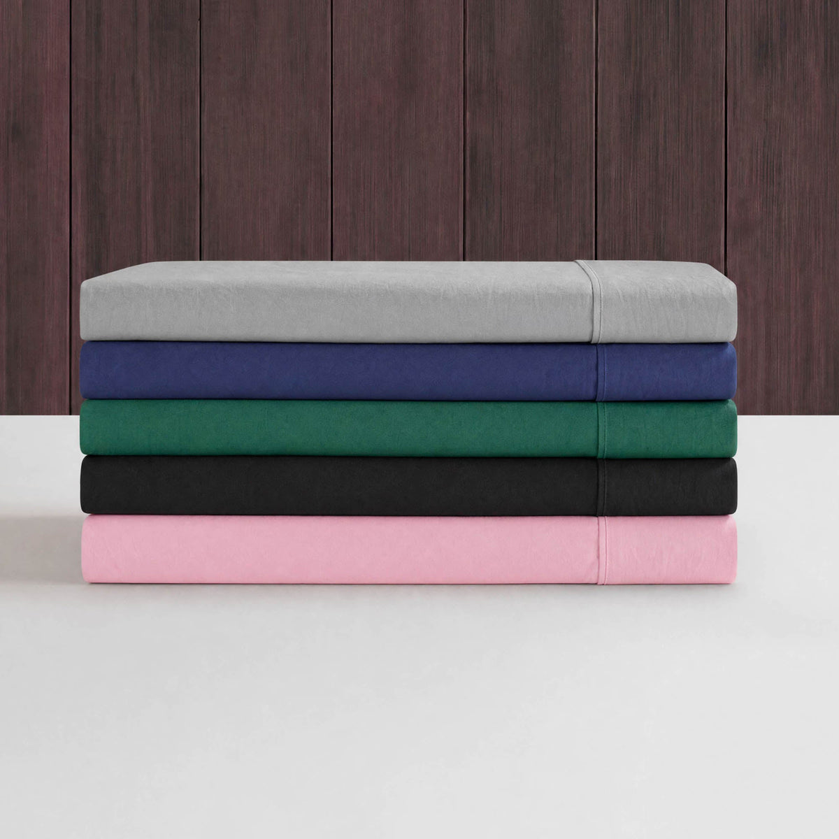 A stack of five neatly folded bedsheets in gray, blue, green, black, and pink—part of the Peak Comfort Microfiber Sheet Set by Champion—is placed on a white surface with a dark wooden background. These athletic bedding deep pocket sheets provide both stylish color choices and a comfortable fit for your bed.