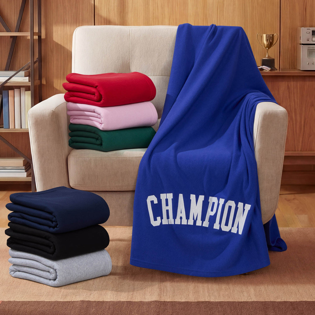 A beige armchair holds a blue Champion Jersey with Fleece Backing Throw Blanket featuring “CHAMPION” text. Extra folded Champion Jersey with Fleece Backing Throw Blankets in pink, red, green, navy, black, and gray are stacked nearby in a wood-paneled room with a trophy on a shelf.