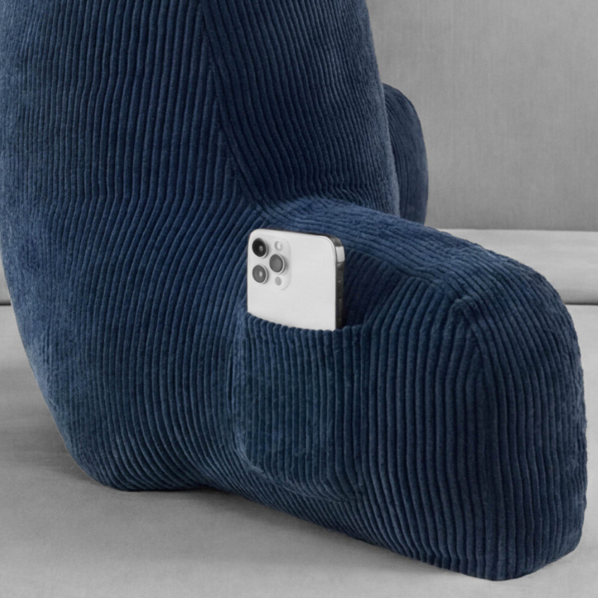 A silver smartphone is placed in the built-in side pocket of a blue Champion Corduroy Backrest Pillow by Champion on a light gray surface.