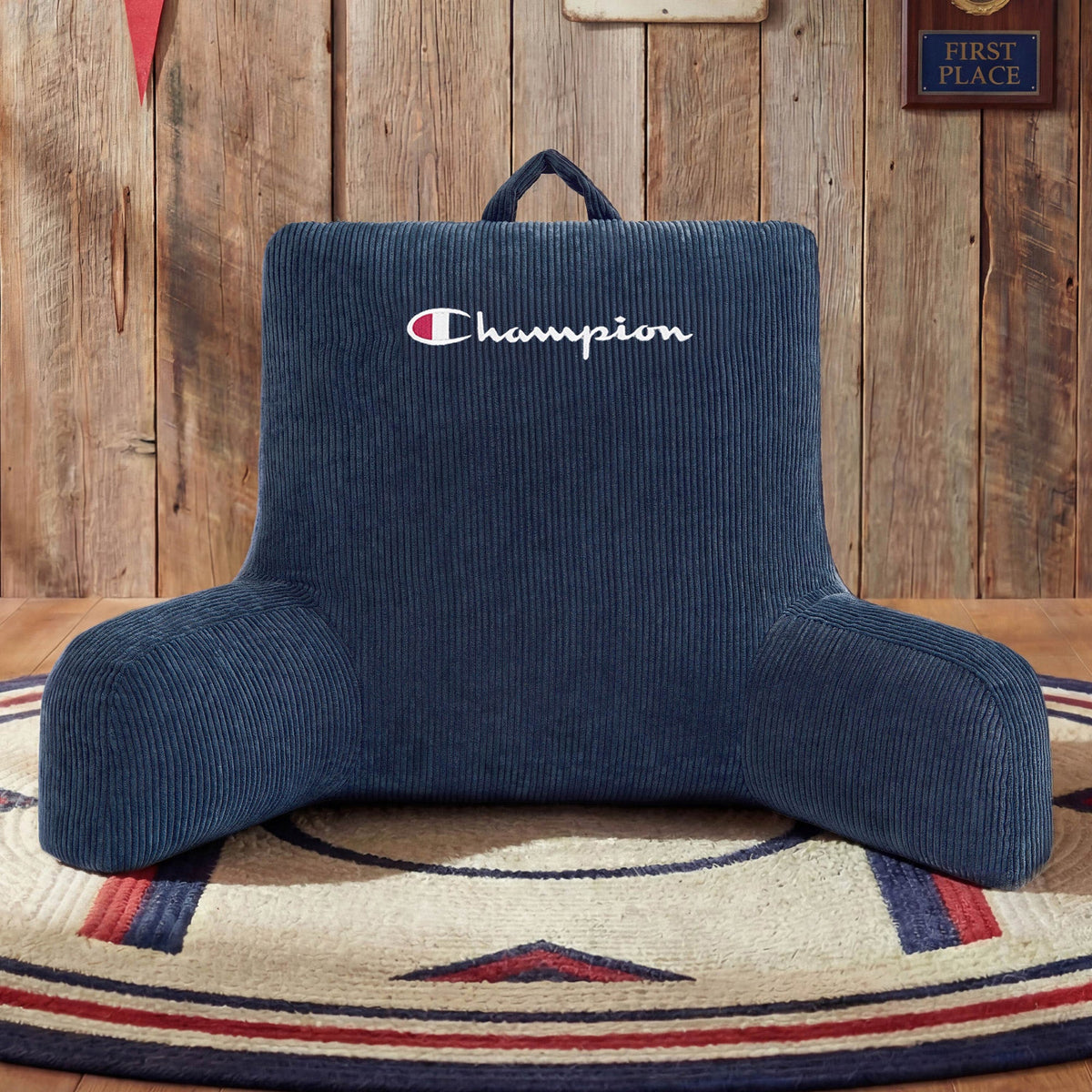 A Champion Corduroy Backrest Pillow in blue, featuring a built-in side pocket, rests on a circular rug with a wood-paneled wall and a First Place sign in the background.