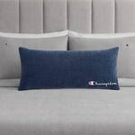 A Champion Corduroy Body Pillow in blue, featuring an embroidered logo, rests on a neatly made bed with light grey sheets, two grey pillows, and a padded grey headboard—creating the perfect lounge space to experience Champion comfort.