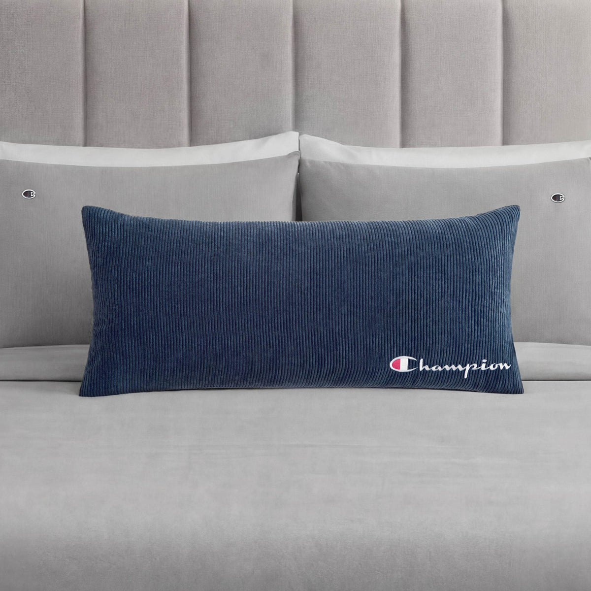 A Champion Corduroy Body Pillow in blue, featuring an embroidered logo, rests on a neatly made bed with light grey sheets, two grey pillows, and a padded grey headboard—creating the perfect lounge space to experience Champion comfort.