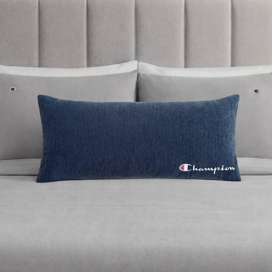 A Champion Corduroy Body Pillow in blue, featuring an embroidered logo, rests on a neatly made bed with light grey sheets, two grey pillows, and a padded grey headboard—creating the perfect lounge space to experience Champion comfort.