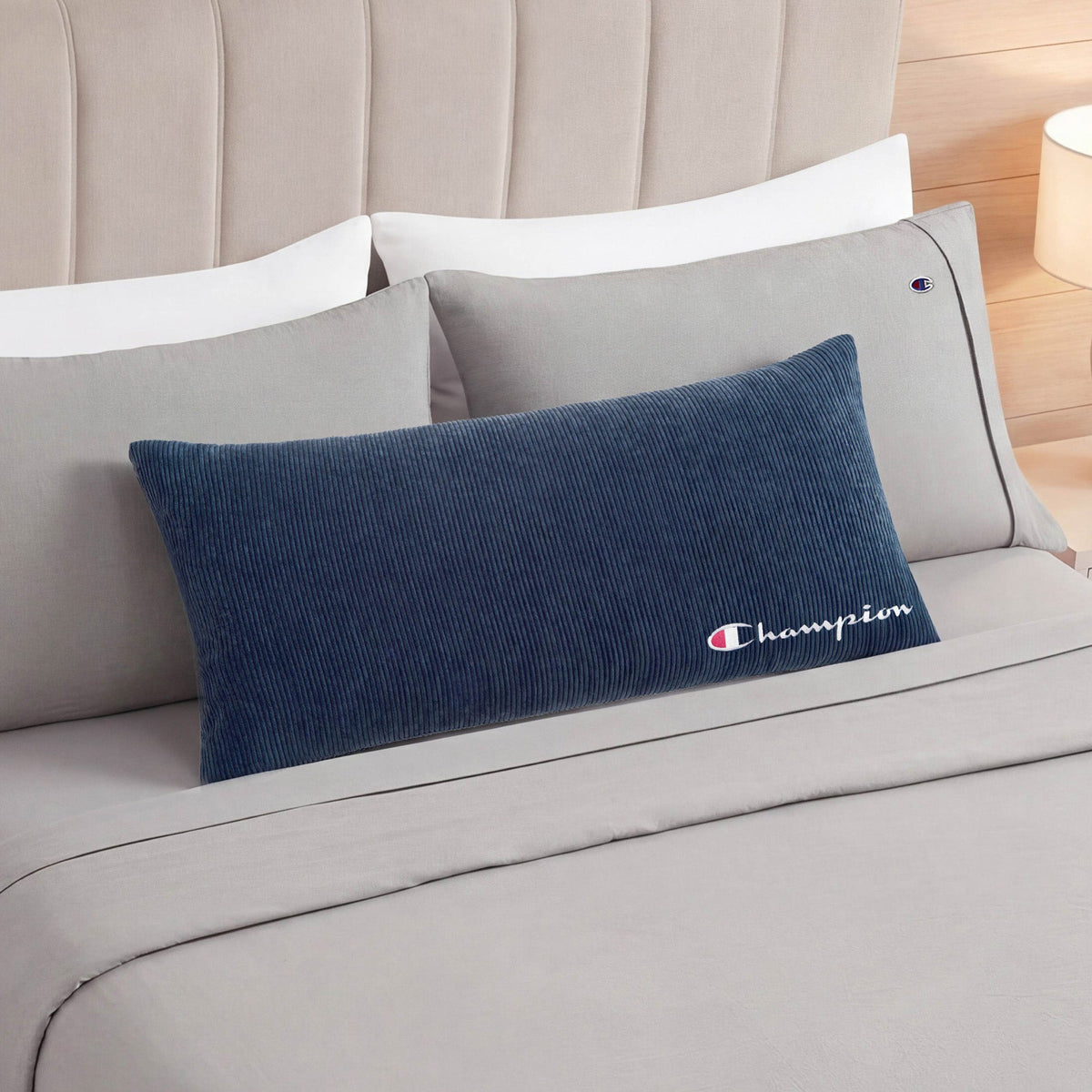 A Champion Corduroy Body Pillow in blue, featuring the Champion logo, rests on a neatly made bed with light gray sheets, gray pillowcases, and a beige upholstered headboard, adding signature Champion comfort to your lounge space.