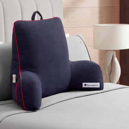 A Champion Faux Wool Backrest Pillow in dark gray, accented with red piping and featuring the small Champion logo, sits on a neatly made bed in a modern bedroom. Crafted with cozy faux wool fabric for added comfort, it is perfect as a bedrest pillow. A white lamp rests on a nearby bedside table.