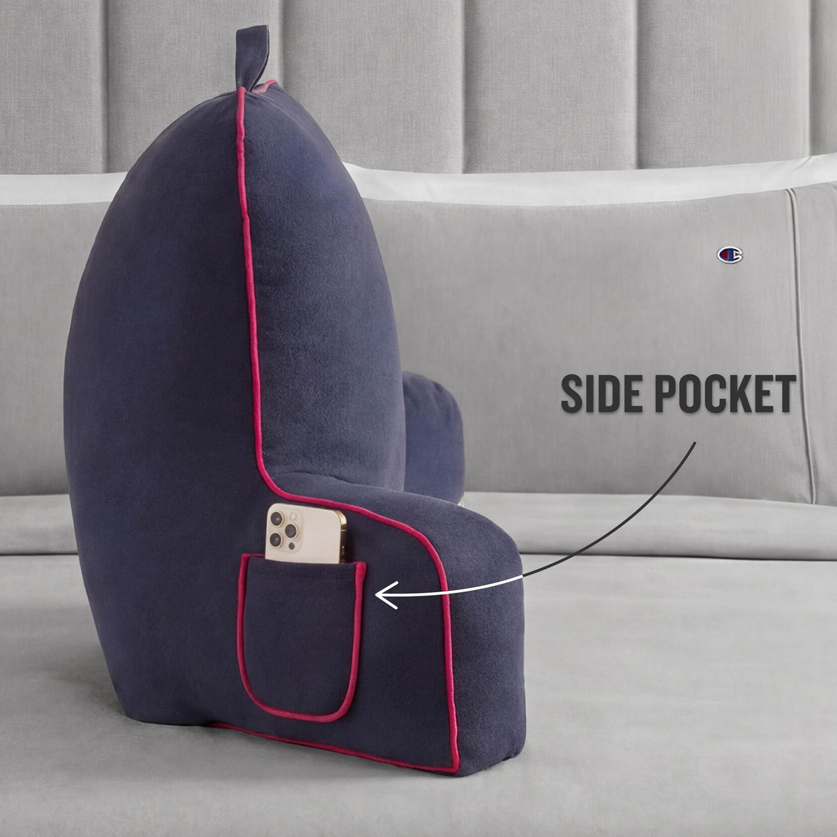 A navy Champion Faux Wool Backrest Pillow with red trim rests on a bed. The backrest pillow by Champion features a side pocket that holds a smartphone, highlighted by an arrow labeled SIDE POCKET.