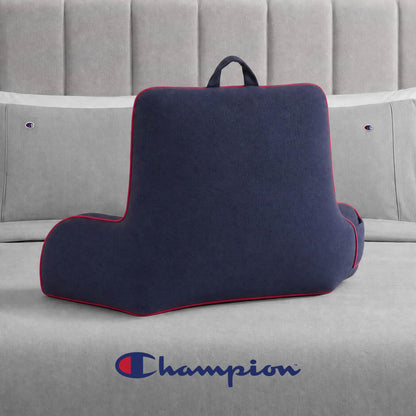 A Champion Faux Wool Backrest Pillow in navy with red trim sits on a gray bed, accompanied by pillows and a headboard featuring the Champion logo. The Champion logo is also visible at the foot of the bed.