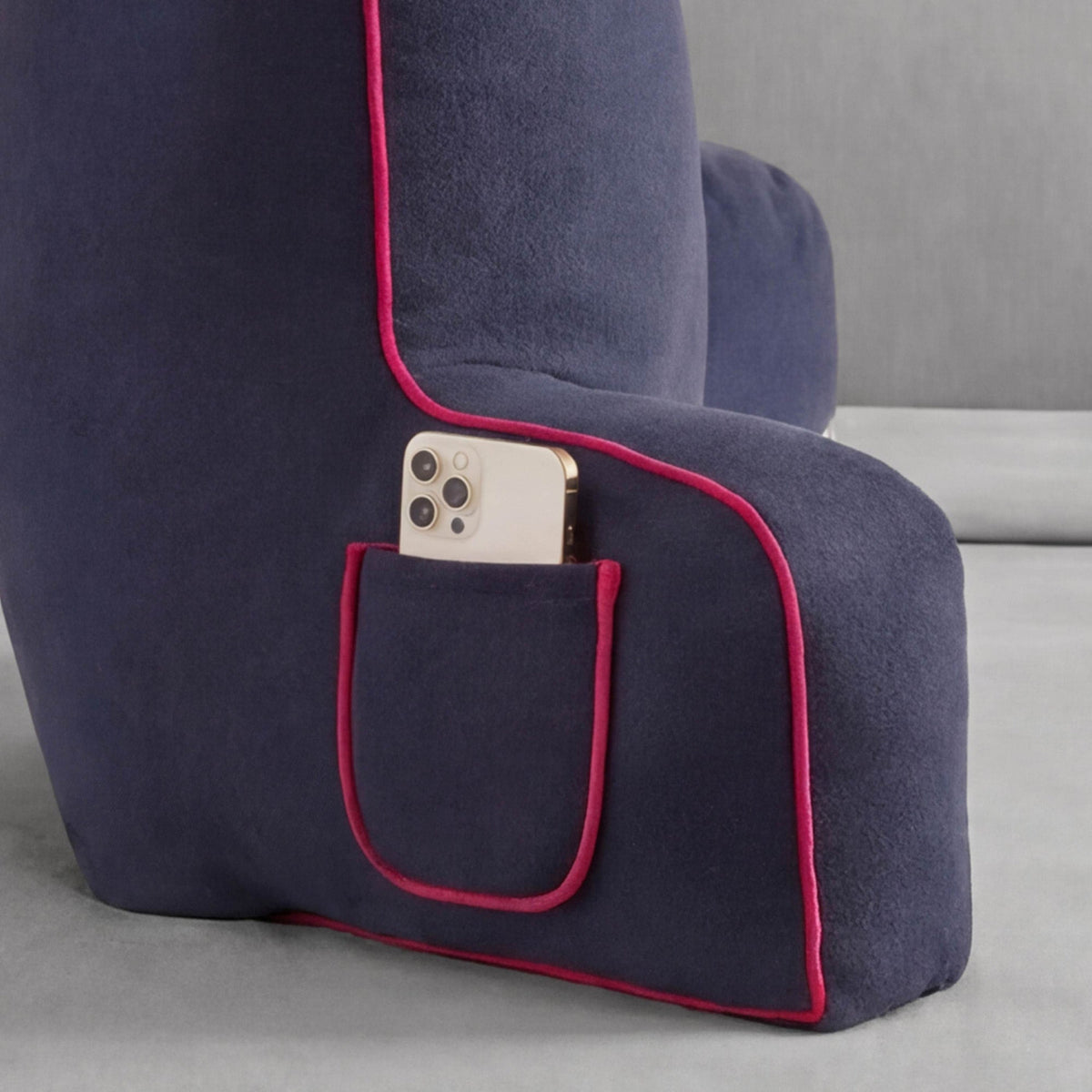 A Champion Faux Wool Backrest Pillow by Champion in dark blue with red piping rests on a light gray surface. A smartphone with three camera lenses is partially visible in a small side pocket of this stylish bedrest pillow.