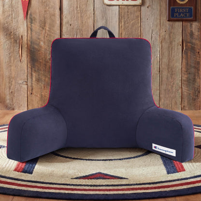A Champion Faux Wool Backrest Pillow in navy blue rests on a round rug in a rustic room with wooden walls and vintage decor. Featuring the Champion logo on the bottom right corner, this cozy bedrest pillow adds classic style and comfort to the space.