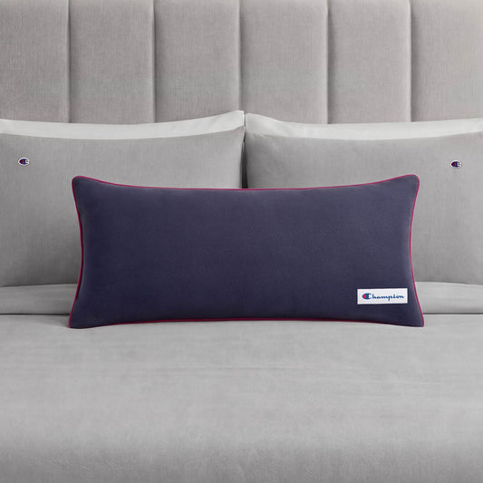 A Champion Faux Wool Body Pillow in navy blue, featuring a small Champion logo, is placed on a neatly made bed with grey sheets and two matching grey pillows, each adorned with a subtle Champion accent.