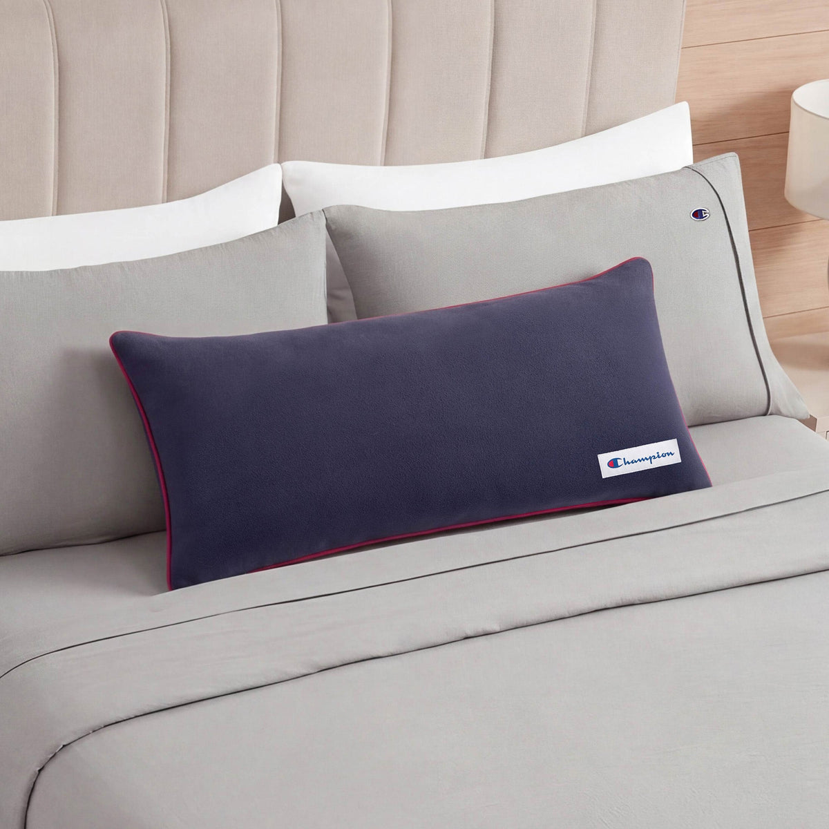 A Champion Faux Wool Body Pillow in navy blue rests on a made bed with light gray sheets, matching light gray pillows, and a beige upholstered headboard.