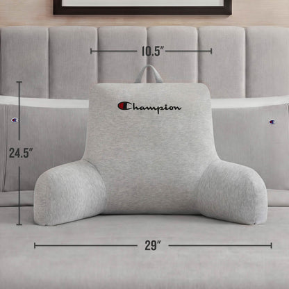 A Champion Jersey Backrest Pillow in gray from Champion rests comfortably on a bed, its arms providing support. The dimensions—24.5 inches tall, 29 inches wide, and 10.5 inches deep—are clearly noted. This cozy pillow is adorned with the iconic Champion logo and placed against a tufted headboard.