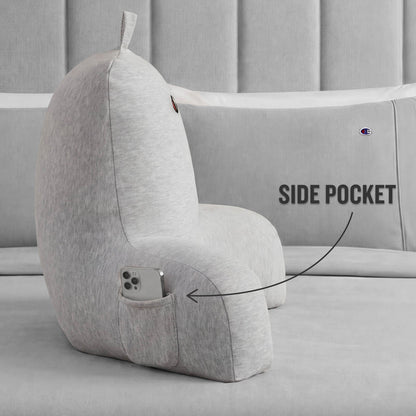 Champion Champion Jersey Backrest Pillow with arms in gray displayed on a bed, featuring a convenient small side pocket holding a smartphone. An arrow and label highlight the pocket design for easy access, with cozy bedding visible in the background.