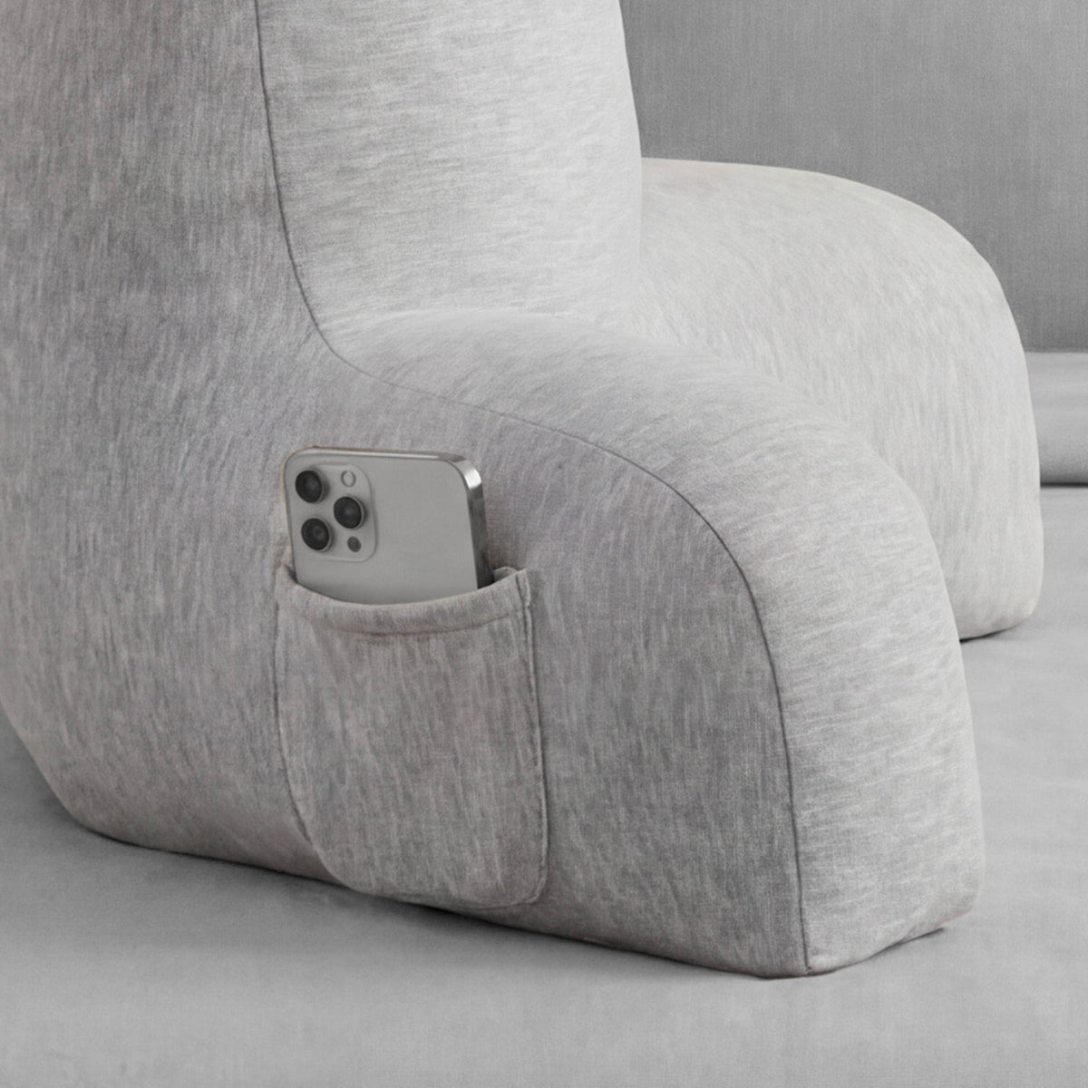 A close-up of the Champion Jersey Backrest Pillow in gray, featuring a small pocket on the armrest that holds a silver smartphone. The gray background blends seamlessly with the pillow, creating a comfortable and signature Champion look.