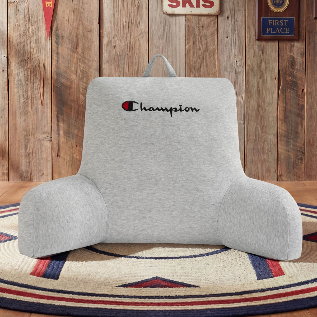 A cozy Champion Jersey Backrest Pillow in gray, featuring the Champion logo embroidery, rests on a round rug with a star motif. The background displays wooden walls decorated with vintage signs and awards.