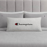 A Champion Jersey Body Pillow in gray, featuring the iconic Champion logo and branding, rests on a coordinating gray bed with two matching pillows and a padded headboard. Made by Champion from soft jersey-knit polyester, this body pillow offers full-length cushioning and comfort for your sleep space.