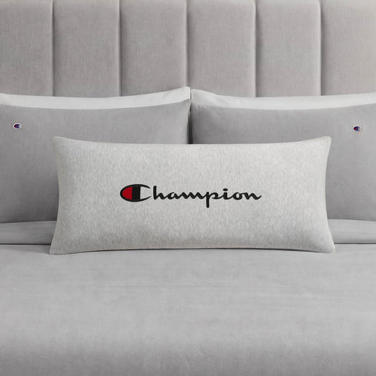 A Champion Jersey Body Pillow in gray, featuring the iconic Champion logo and branding, rests on a coordinating gray bed with two matching pillows and a padded headboard. Made by Champion from soft jersey-knit polyester, this body pillow offers full-length cushioning and comfort for your sleep space.