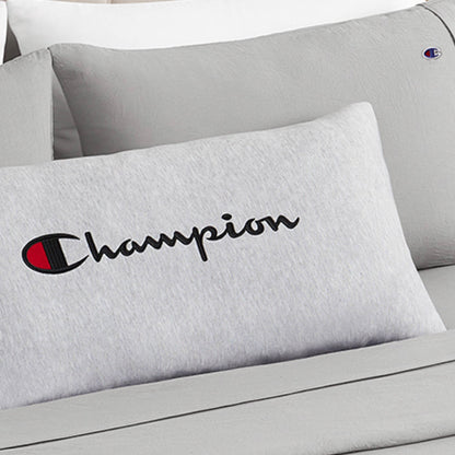 A close-up of a Champion Jersey Body Pillow in gray jersey-knit polyester, showcasing the iconic Champion logo and text, rests on a matching gray bed alongside another pillow featuring the brand’s emblem. This cozy setup, with its full-length cushioning, is perfect for relaxing with your favorite Champion body pillow.