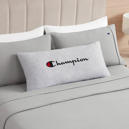 A gray rectangular Champion Jersey Body Pillow from Champion, made of soft jersey-knit polyester for full-length cushioning, is placed on a neatly made bed with light gray sheets and pillows, set against a light-colored upholstered headboard.