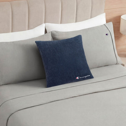 A neatly made bed with light gray sheets and pillows features a Champion 18" x 18" Logo Pillow in dark blue corduroy, showcasing the brand’s iconic logo and adding a casual style touch against the pillows. A beige headboard and a small bedside lamp are visible.