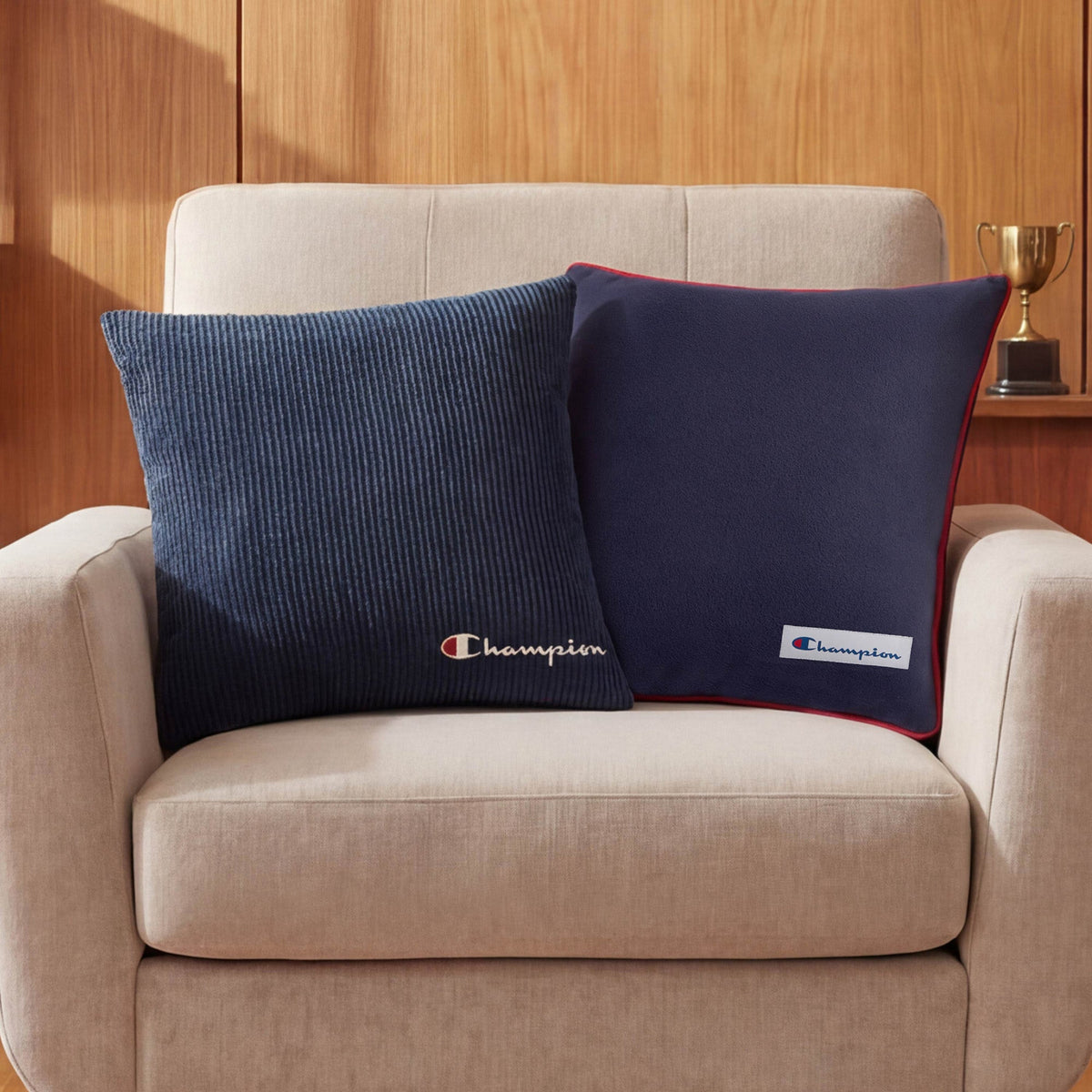 A beige armchair with two Champion 18" x 18" Logo Pillows, each featuring a soft polyester cover in navy blue, sits in front of a wooden wall and shelf with a gold trophy.