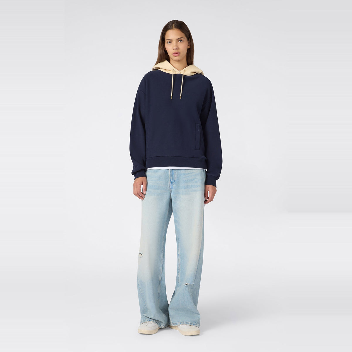 A person stands facing forward, wearing a navy Champion Women’s Knitted Hooded Sweatshirt layered over a beige hood, paired with light blue wide-leg jeans and white sneakers, against a plain light gray background.