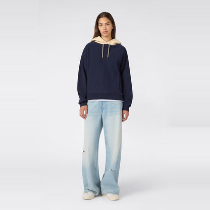 A person stands facing forward, wearing a navy Champion Women’s Knitted Hooded Sweatshirt layered over a beige hood, paired with light blue wide-leg jeans and white sneakers, against a plain light gray background.