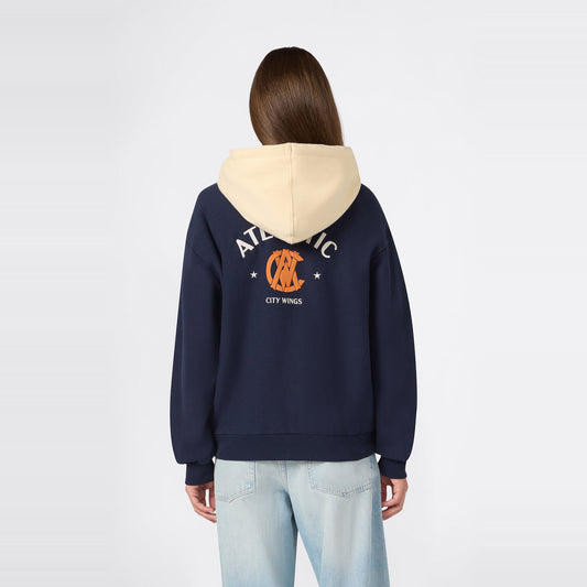 A person wearing the Champion Women’s Knitted Hooded Sweatshirt in navy, featuring a beige hood, “ATLANTIC” text, an orange graphic on the front, and “CITY WINGS” printed on the back, paired with light blue jeans, stands facing away from the camera against a plain background.