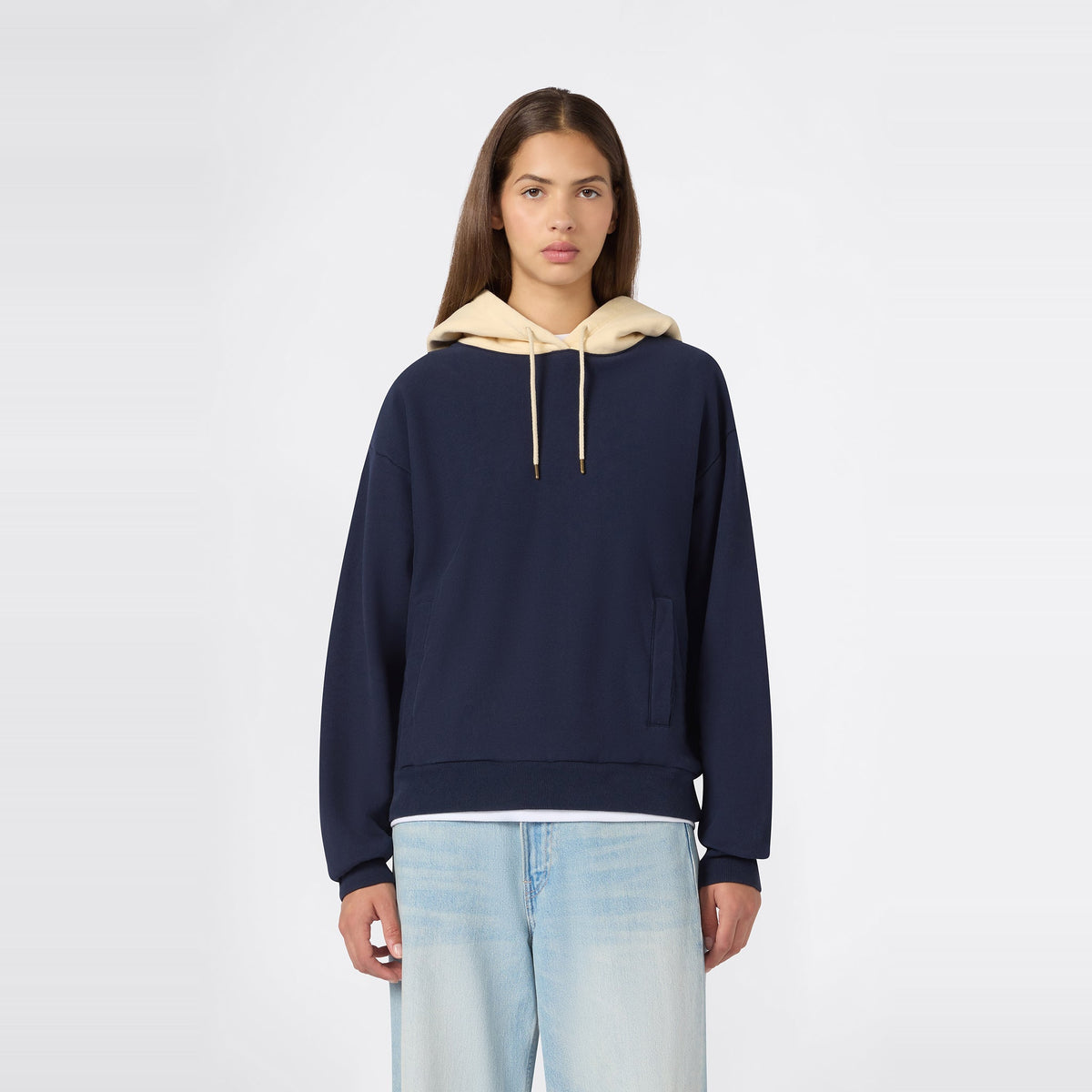 A person wearing the Champion Women’s Knitted Hooded Sweatshirt in navy blue, featuring a beige hood and heavyweight fleece construction, paired with light blue jeans, stands facing forward against a plain white background.
