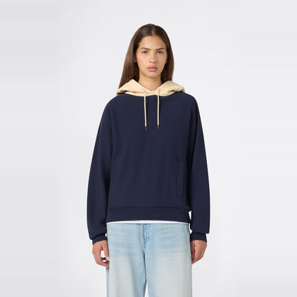 A person wearing the Champion Women’s Knitted Hooded Sweatshirt in navy blue, featuring a beige hood and heavyweight fleece construction, paired with light blue jeans, stands facing forward against a plain white background.