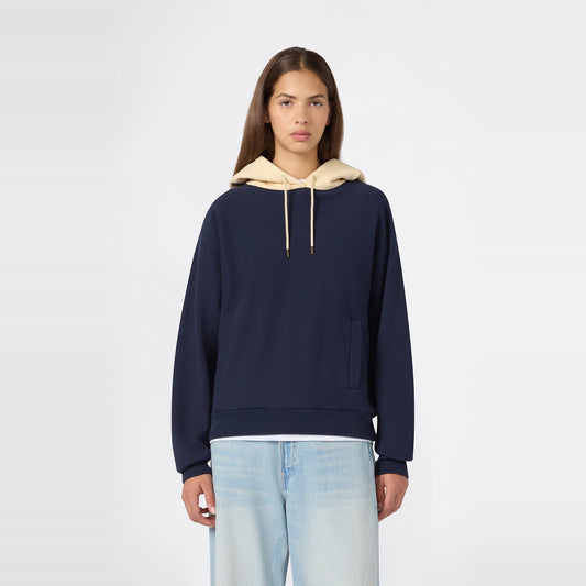 A person wearing the Champion Women’s Knitted Hooded Sweatshirt in navy blue, featuring a beige hood and heavyweight fleece construction, paired with light blue jeans, stands facing forward against a plain white background.
