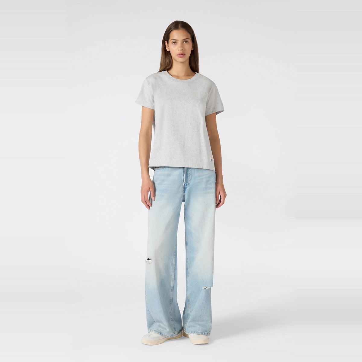 A woman stands facing forward against a plain white background, wearing the Champion Women’s Knitted Crewneck T-Shirt in grey with a relaxed fit, paired with light blue wide-leg jeans featuring subtle distressing and white sneakers.