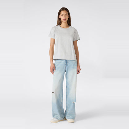 A woman stands facing forward against a plain white background, wearing the Champion Women’s Knitted Crewneck T-Shirt in grey with a relaxed fit, paired with light blue wide-leg jeans featuring subtle distressing and white sneakers.