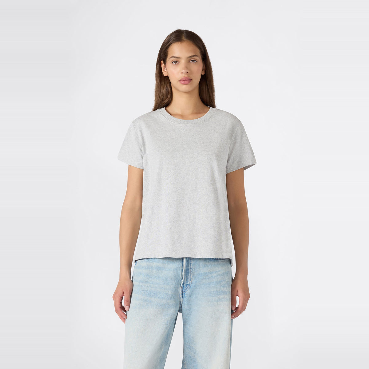 A person with long brown hair stands facing forward against a plain white background, wearing a relaxed fit Champion Women’s Knitted Crewneck T-Shirt in plain light gray paired with light blue jeans.