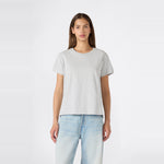 A person with long brown hair stands facing forward against a plain white background, wearing a relaxed fit Champion Women’s Knitted Crewneck T-Shirt in plain light gray paired with light blue jeans.