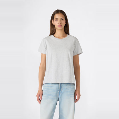 A person with long brown hair stands facing forward against a plain white background, wearing a relaxed fit Champion Women’s Knitted Crewneck T-Shirt in plain light gray paired with light blue jeans.