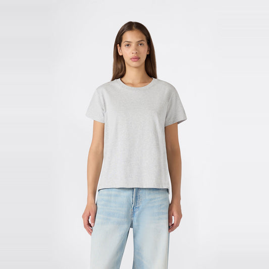 A person with long brown hair stands facing forward against a plain white background, wearing a relaxed fit Champion Women’s Knitted Crewneck T-Shirt in plain light gray paired with light blue jeans.