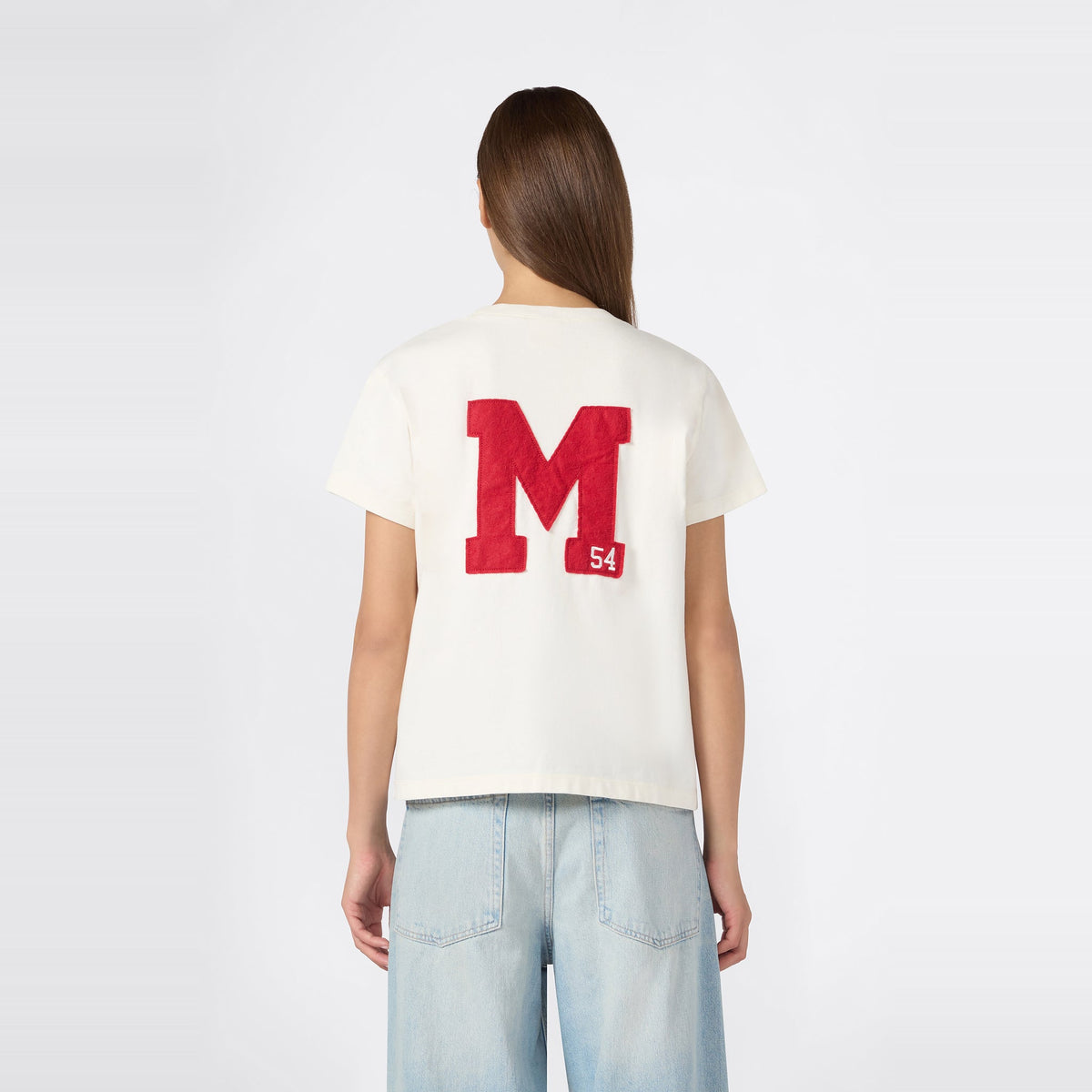 A woman with long brown hair is wearing the Champion Women’s Knitted Crewneck T-Shirt in white, featuring a bold red “M” and a small “54” on the back, paired with relaxed fit light blue jeans, standing against a plain white background.