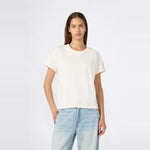 A woman with straight brown hair is wearing a relaxed fit Champion Women’s Knitted Crewneck T-Shirt in plain white paired with light blue jeans, standing against a light gray background with one hand in her pocket.