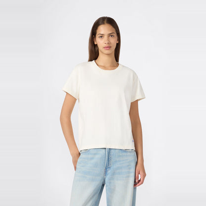A woman with straight brown hair is wearing a relaxed fit Champion Women’s Knitted Crewneck T-Shirt in plain white paired with light blue jeans, standing against a light gray background with one hand in her pocket.