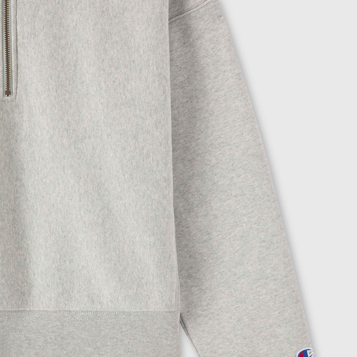 Close-up of a light gray Champion Women’s Knitted Half Zip Sweatshirt with long sleeves. A small patch featuring the blue and red Champion logo is visible on the cuff, highlighting this essential piece of women’s sportswear.