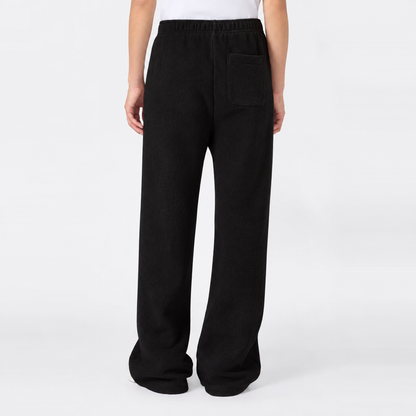 A person is seen from the waist down, standing against a plain white background, wearing Champion Women’s Knitted Flare Pants—loose-fitting black trousers featuring an elastic waistband and back pocket for a modern twist on classic sportswear style.