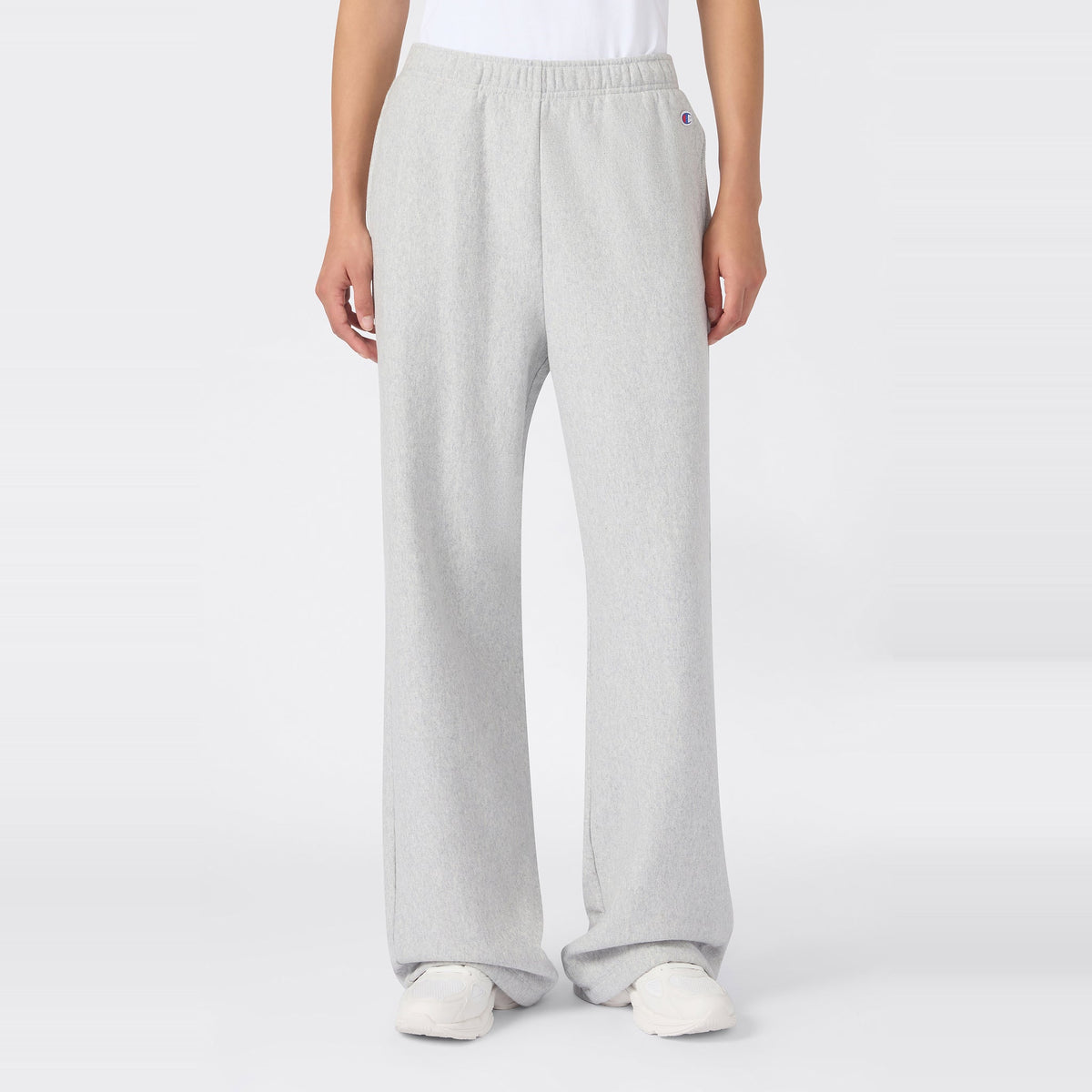A person wearing Champion Women’s Knitted Flare Pants in a loose-fit, light gray style, paired with a white t-shirt and white sneakers, stands against a plain white background. The heavyweight fleece fabric of these flared pants adds an elevated touch to this classic women’s sportswear look.