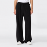 A person wearing loose black Champion Women’s Knitted Flare Pants and white sneakers stands against a plain white background. Only the lower half of the body is visible, with hands relaxed at the sides, highlighting a laid-back approach to women’s sportswear.