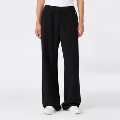 A person wearing loose black Champion Women’s Knitted Flare Pants and white sneakers stands against a plain white background. Only the lower half of the body is visible, with hands relaxed at the sides, highlighting a laid-back approach to women’s sportswear.