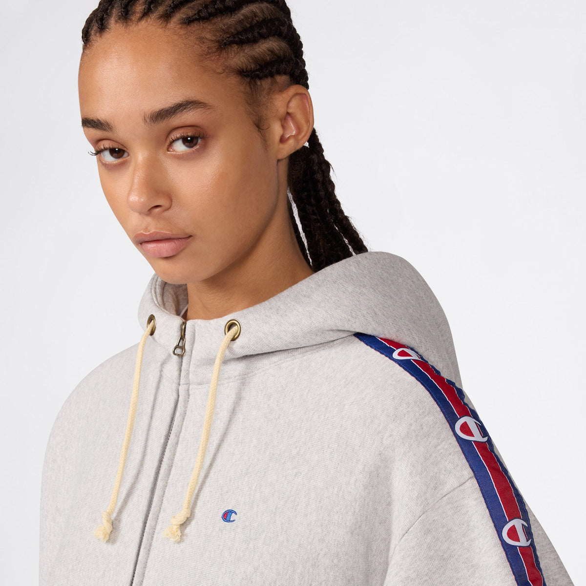 A person with braided hair wears the Women’s Icon Hooded Full Zip Sweatshirt by Champion in light gray, featuring blue and red logo stripes on the sleeves, looking slightly to the side against a plain background. The heavyweight fleece construction of this hoodie provides both comfort and durability, making it a stylish choice.