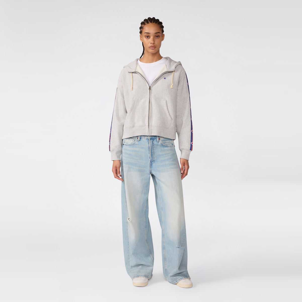 A person stands facing forward, wearing a Champion Women’s Icon Hooded Full Zip Sweatshirt in gray over a white shirt and loose-fitting light blue jeans, paired with white sneakers, against a plain light gray background.