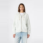 A person stands facing forward, wearing a Champion Women’s Knitted Hooded Full Zip Sweatshirt in a light color over a white shirt and paired with light blue jeans, against a plain white background.